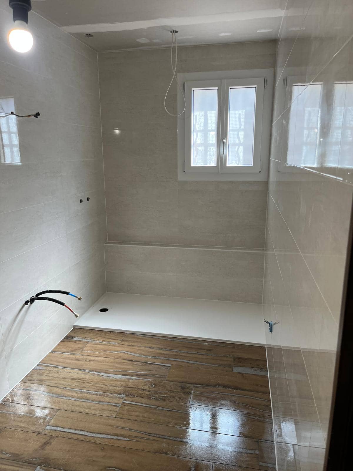 Faïence claire gris-beige posée à l’horizontale, grand receveur extraplat blanc devant un muret carrelé sous la fenêtre, sol en carrelage effet parquet, réalisation à Bourg-en-Bresse
