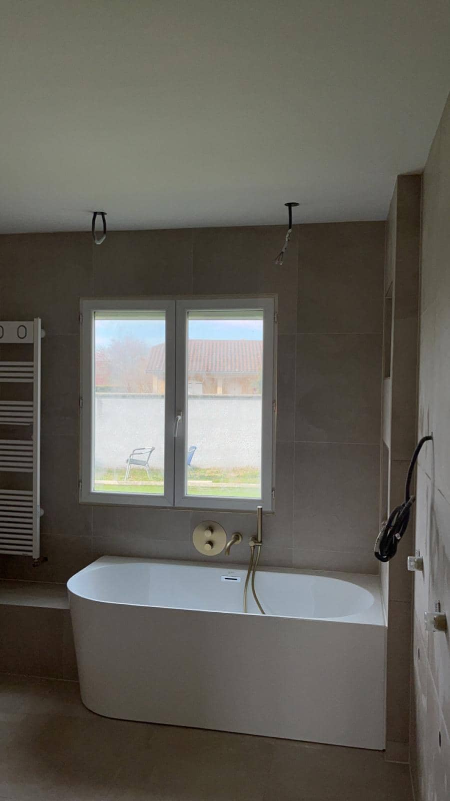Baignoire adossée sous une fenêtre, murs et sol en grands carreaux beige-grège aspect pierre, robinetterie encastrée or brossé, luminaires pas encore posés, réalisation à Gex