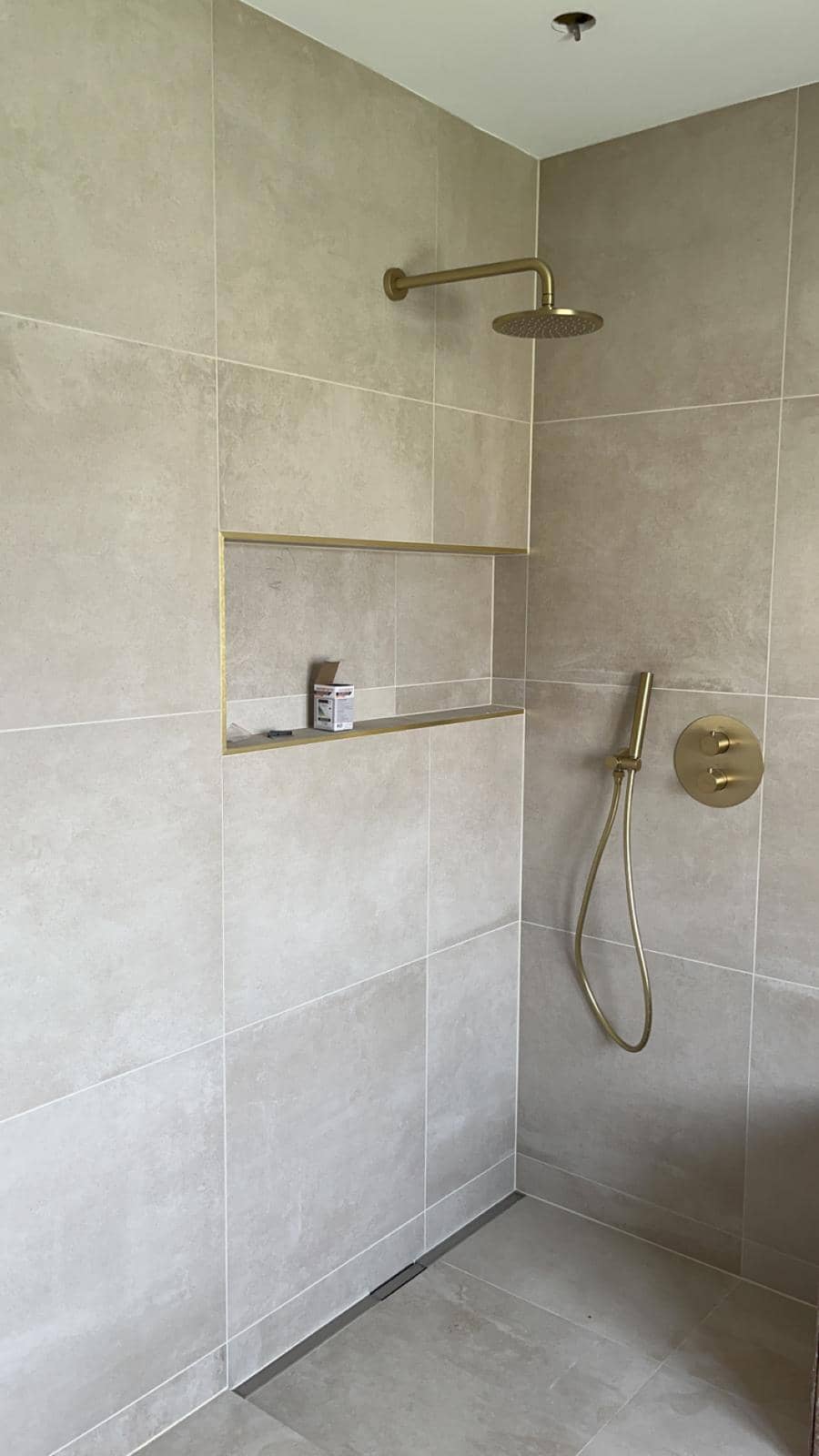 Douche de plain-pied entièrement carrelée en grands carreaux beige aspect pierre, caniveau linéaire en pied de mur, niche horizontale à profilé doré et robinetterie or brossé, réalisation à Gex