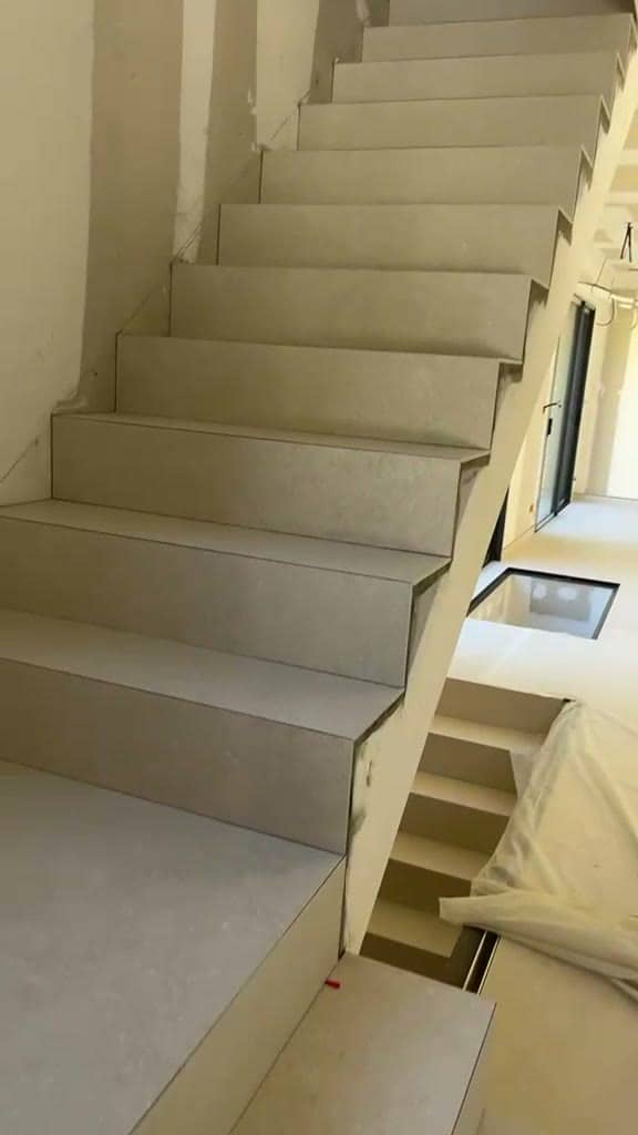Volée d’escalier carrelée en grès beige clair, départ d’une seconde volée et panneau vitré au sol du palier, murs encore bruts, réalisation à Divonne-les-Bains