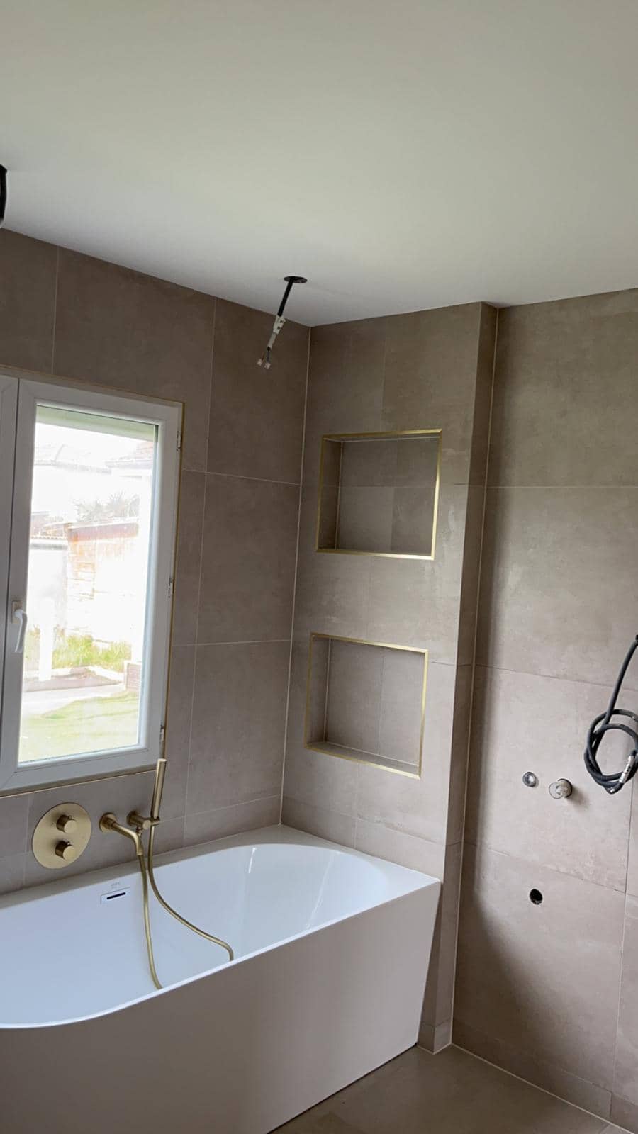 Deux niches superposées cernées de profilés dorés au-dessus d’une baignoire adossée, murs en grands carreaux beige aspect pierre à joints fins clairs, réalisation à Gex