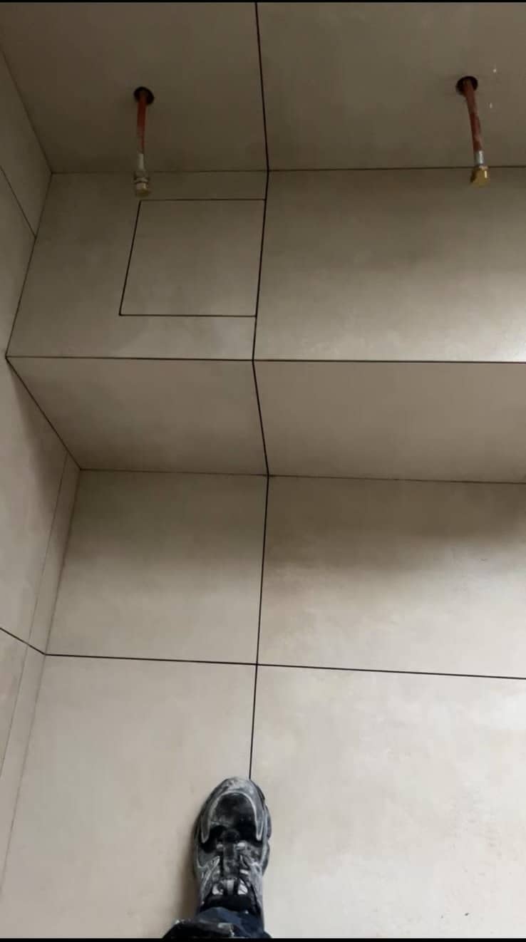 Vue plongeante sur un sol et un podium en grands carreaux beige à joints fins foncés alignés, trappe de visite intégrée, chaussure d’ouvrier au premier plan, réalisation à Chambéry