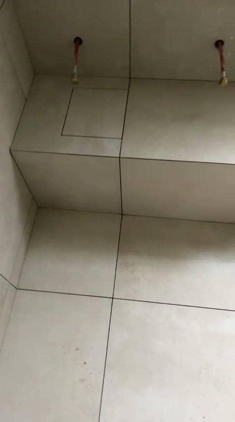 Podium carrelé en grands carreaux beige avec trappe de visite intégrée sur le dessus, joints foncés filant du sol jusqu’au mur, deux arrivées d’eau en attente, réalisation à Chambéry