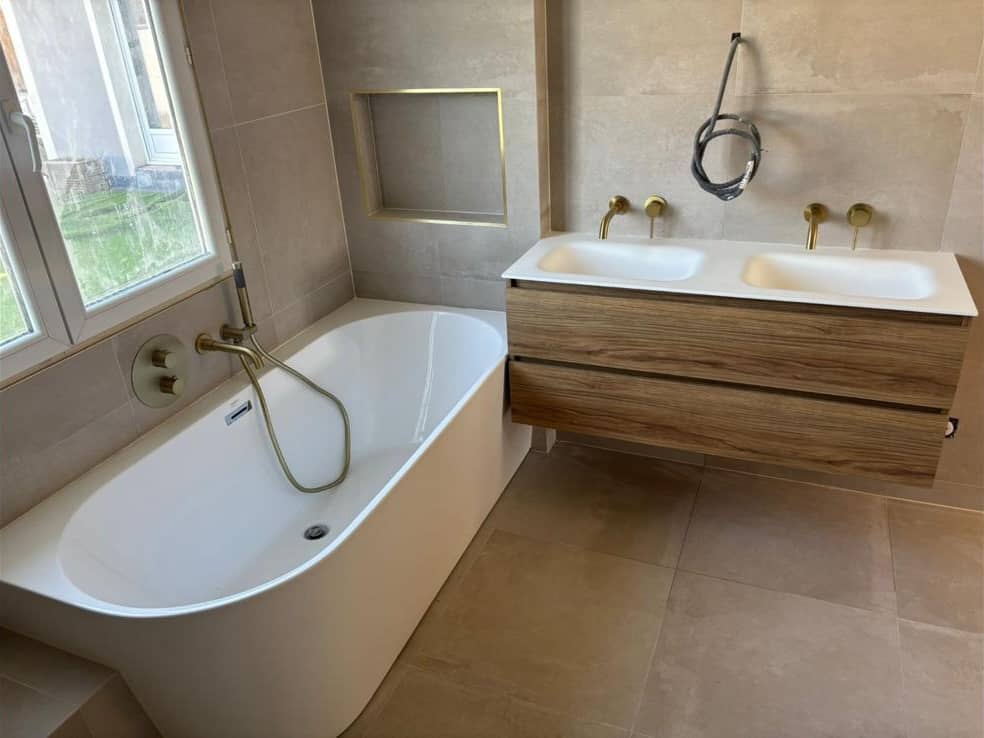 Salle de bain terminée en grès cérame beige aspect pierre grand format au sol et aux murs, baignoire adossée, niche à profilé doré et meuble double vasque en bois, réalisation à Gex