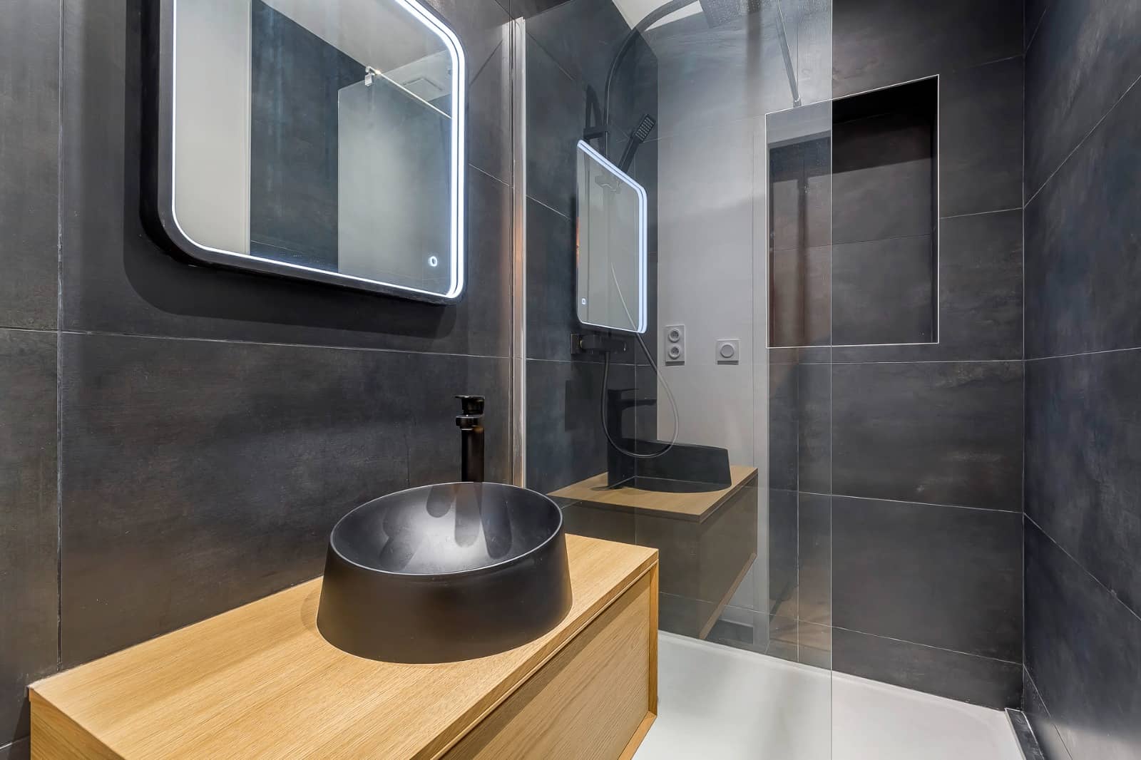 Salle de douche terminée, faïence anthracite grand format posée verticalement, niche carrelée derrière la paroi vitrée, vasque noire mate sur meuble chêne et miroir rétroéclairé, réalisation à Annemasse