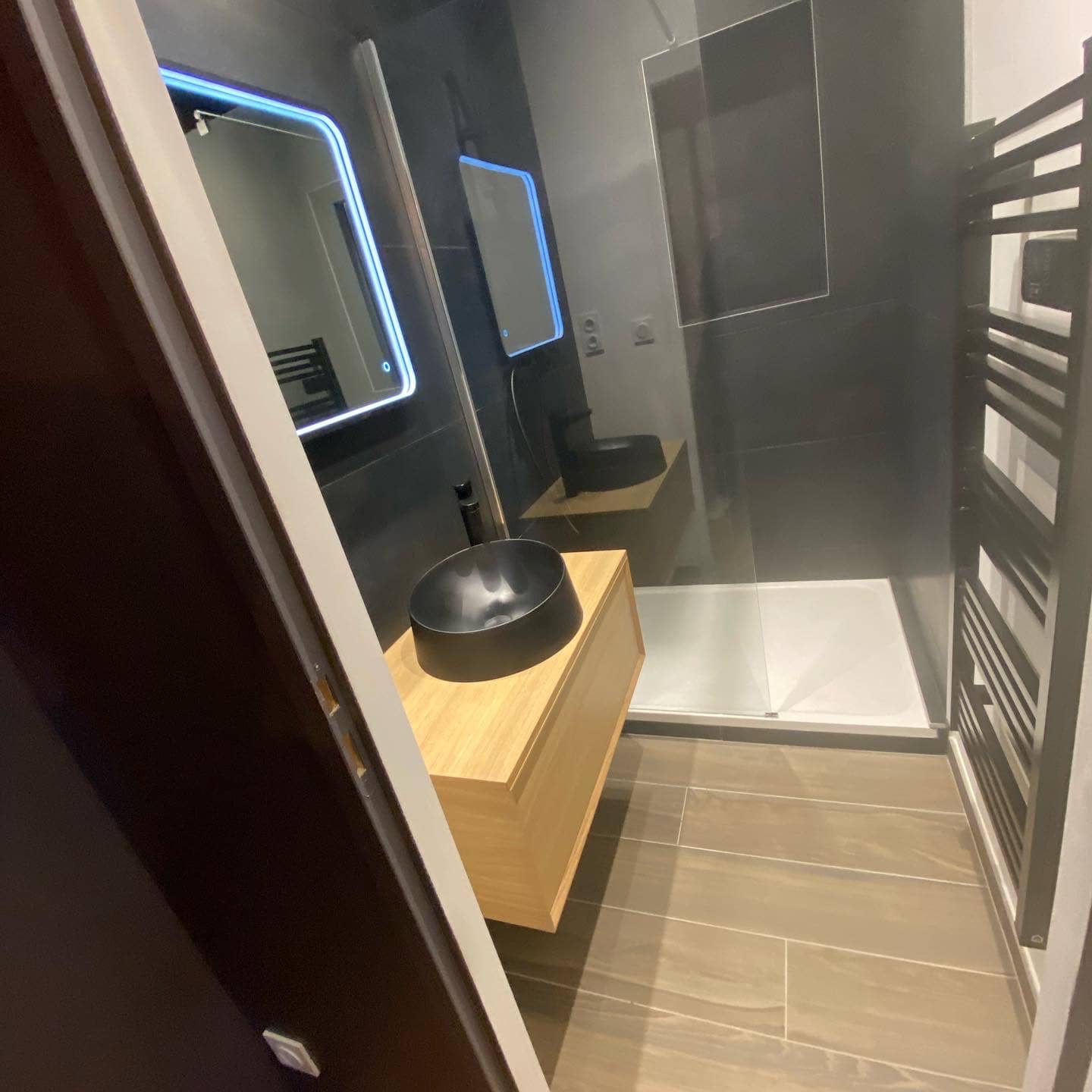 Salle de douche vue depuis la porte, sol en carrelage imitation bois, faïence anthracite, vasque noire sur meuble chêne et miroir rétroéclairé allumé, réalisation à Annemasse