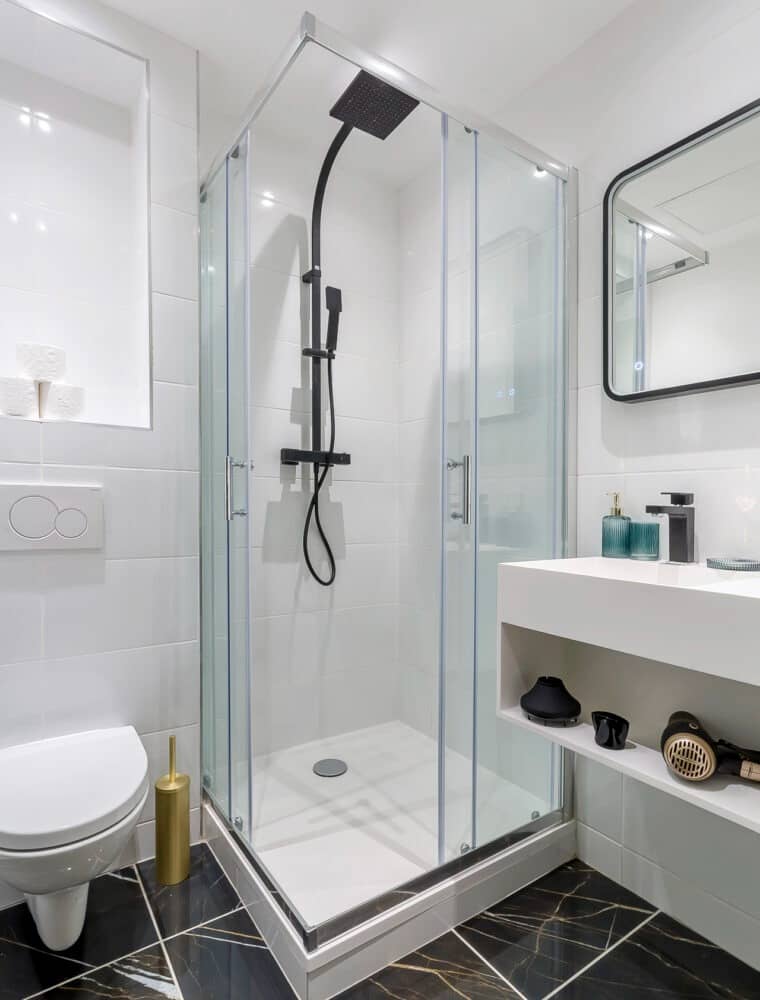 Petite salle de douche terminée, sol effet marbre noir veiné doré posé en diagonale, faïence blanche brillante, cabine d’angle vitrée et WC suspendu, réalisation à Bourg-Saint-Maurice