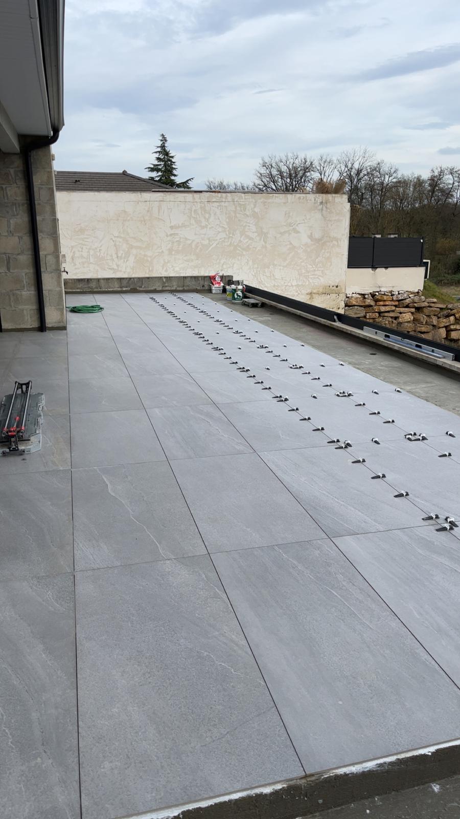 Pose de grandes dalles de grès cérame gris effet pierre sur une terrasse, clips et cales de nivellement sur les derniers rangs, coupe-carreau manuel au premier plan, réalisation à Rumilly