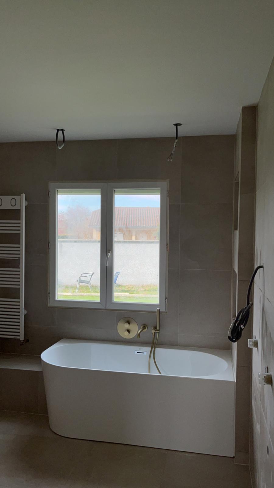 Baignoire adossée sous une fenêtre, murs et sol en grands carreaux beige-grège aspect pierre, robinetterie encastrée or brossé, luminaires pas encore posés, réalisation à Gex