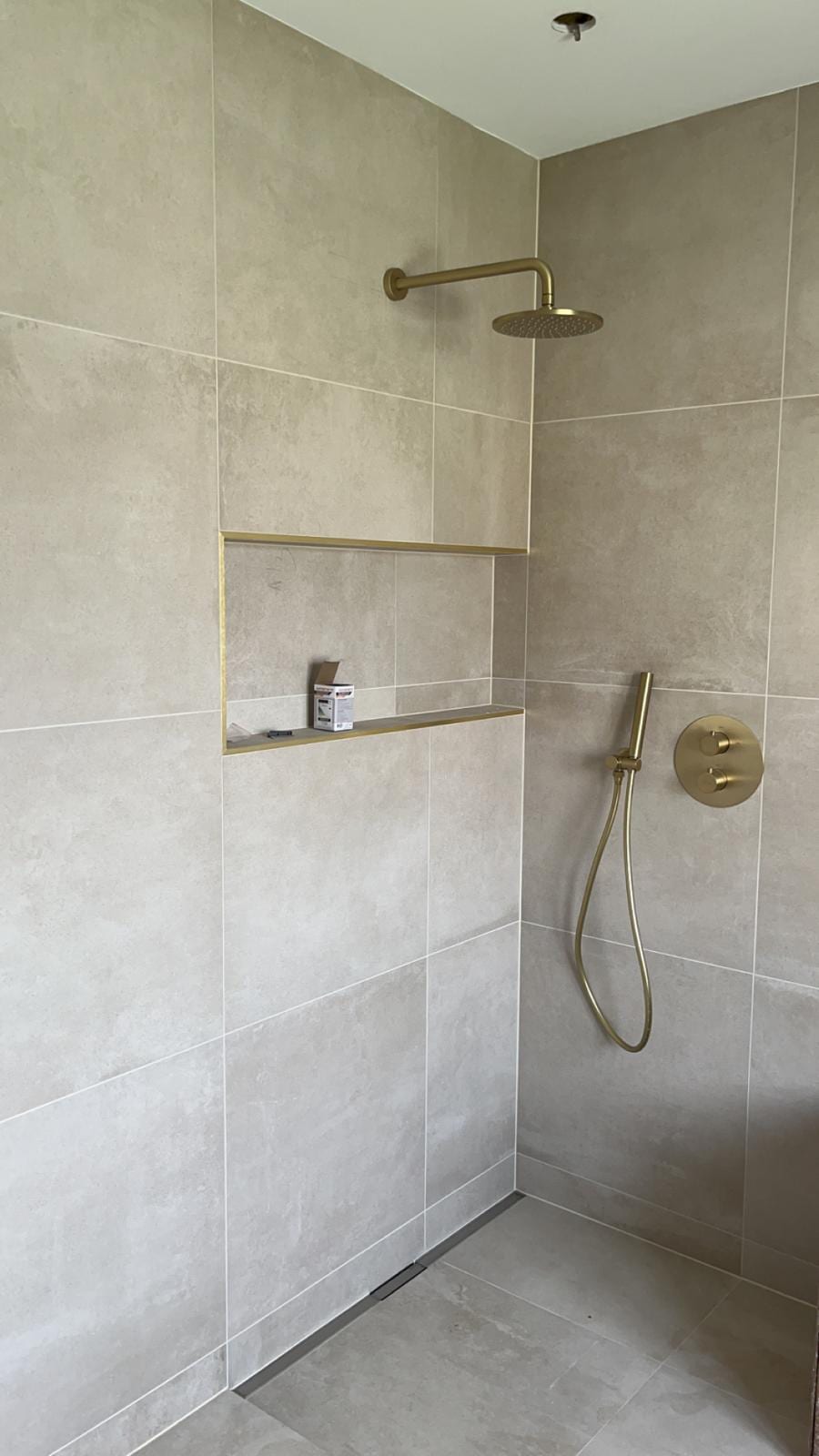 Douche de plain-pied entièrement carrelée en grands carreaux beige aspect pierre, caniveau linéaire en pied de mur, niche horizontale à profilé doré et robinetterie or brossé, réalisation à Gex