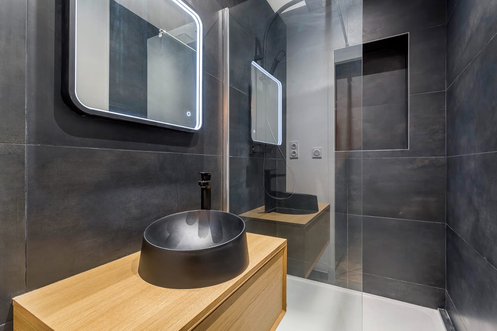 Salle de douche terminée, faïence anthracite grand format posée verticalement, niche carrelée derrière la paroi vitrée, vasque noire mate sur meuble chêne et miroir rétroéclairé, réalisation à Annemasse