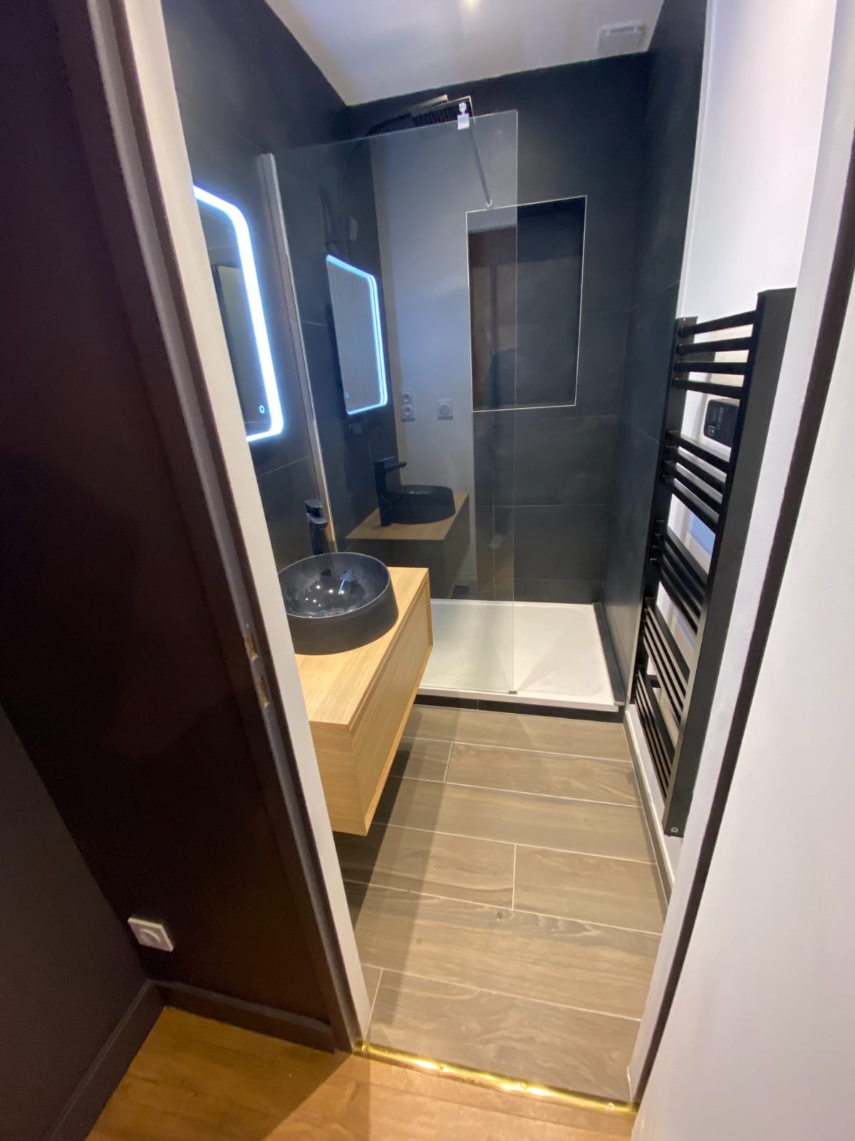 Sol en carrelage imitation bois à joints clairs, barre de seuil laiton vers le parquet du couloir, faïence anthracite et paroi de douche vitrée, réalisation à Annemasse