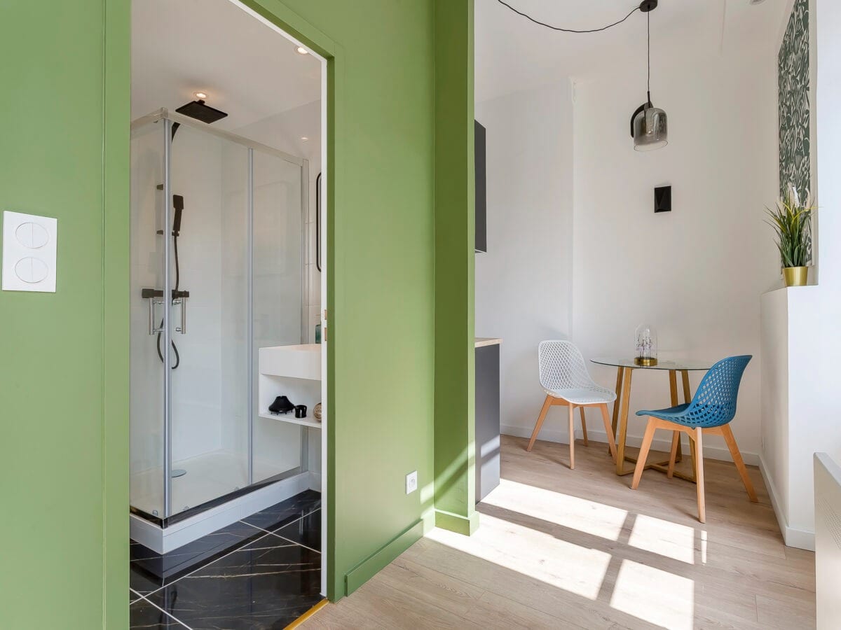 Salle d’eau d’un studio ouverte par une cloison vert olive, cabine de douche vitrée et sol en carrelage effet marbre noir veiné, coin repas à droite, réalisation à Bourg-Saint-Maurice