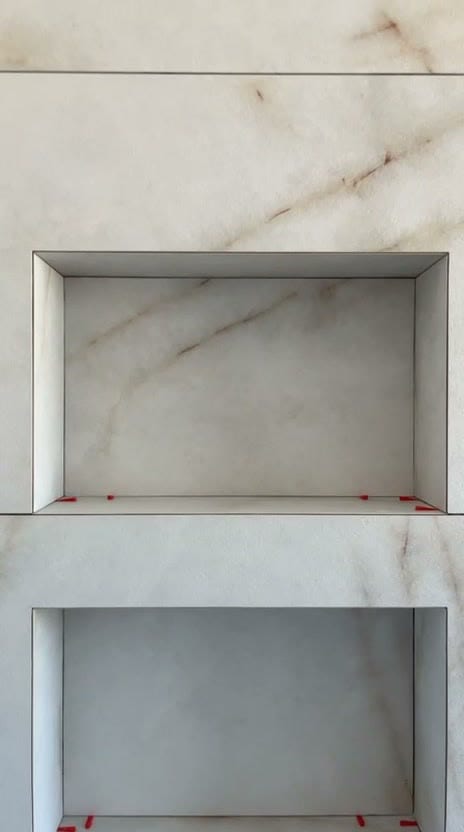 Deux niches superposées habillées de grès effet marbre blanc veiné beige, arêtes nettes sans profilé, cales rouges encore posées sur les tablettes, réalisation à Thonon-les-Bains